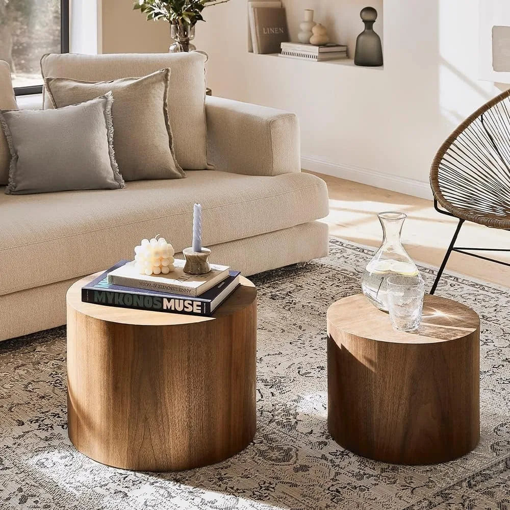 A cozy living room features a beige sofa with tan cushions and a set of two Morsale Living Room Wood Coffee Tables. One table holds books, a candle, and a vase; the other has a glass pitcher. A patterned rug and a woven-back chair complete the furniture ensemble.