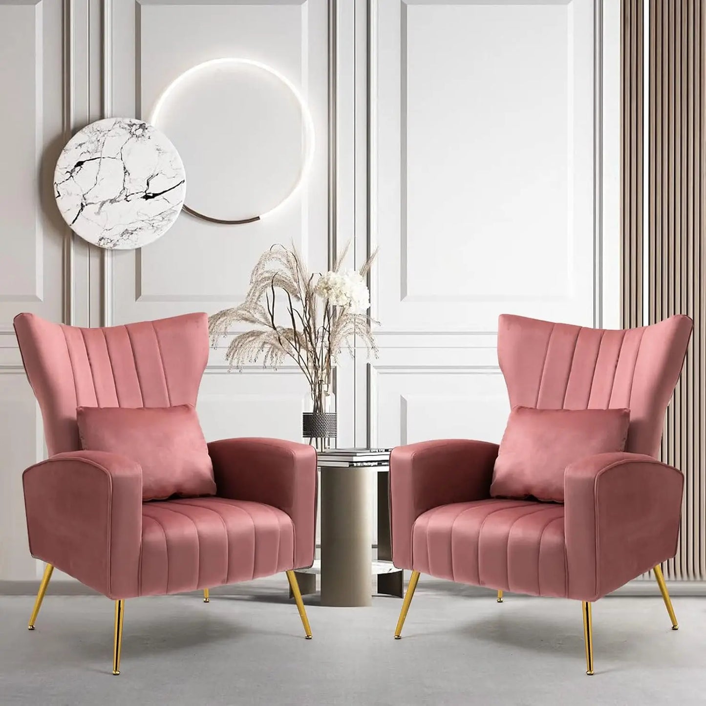 Two Morsale Modern Velvet Lounge Chairs, featuring gold metal legs and a pink hue, are symmetrically placed in a chic room. A small table with a vase of dried flowers sits between them. The wall features vertical lines and a circular art piece with halo lighting above.