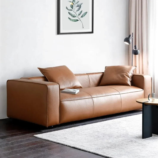 A modern living room showcases the brown Stefano Genuine Leather Lounge Sofa by Morsale, epitomizing modern style. Two matching cushions rest on it with a small book. A framed leaf print adorns the white wall above. A floor lamp complements light curtains, while a black round table and gray rug complete the decor.