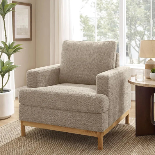 A Morsale Oversized Mid Century Modern armchair in beige, with wooden legs, is placed in a well-lit living room near a large window. The space includes a potted plant, wooden side table holding a lamp and glass of water, framed picture on the wall, and is adorned with a woven rug.