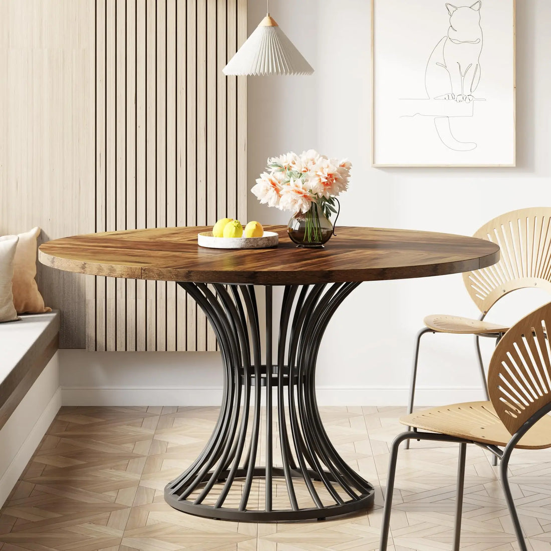 A modern dining area features the Morsale Preston 47-Inch Round Dining Table with an ornate black metal base, adorned with a vase of pink flowers and a bowl of lemons. Two wooden chairs, a wall bench, a pendant lamp, and a cat sketch complete this cozy setting.