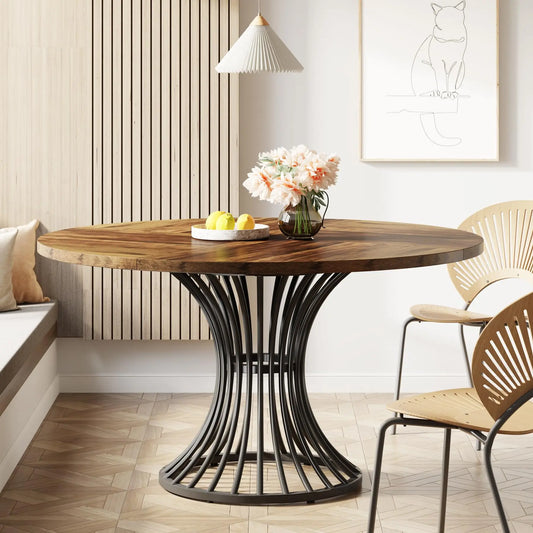 A modern dining area features the Morsale Preston 47-Inch Round Dining Table with an ornate black metal base, adorned with a vase of pink flowers and a bowl of lemons. Two wooden chairs, a wall bench, a pendant lamp, and a cat sketch complete this cozy setting.