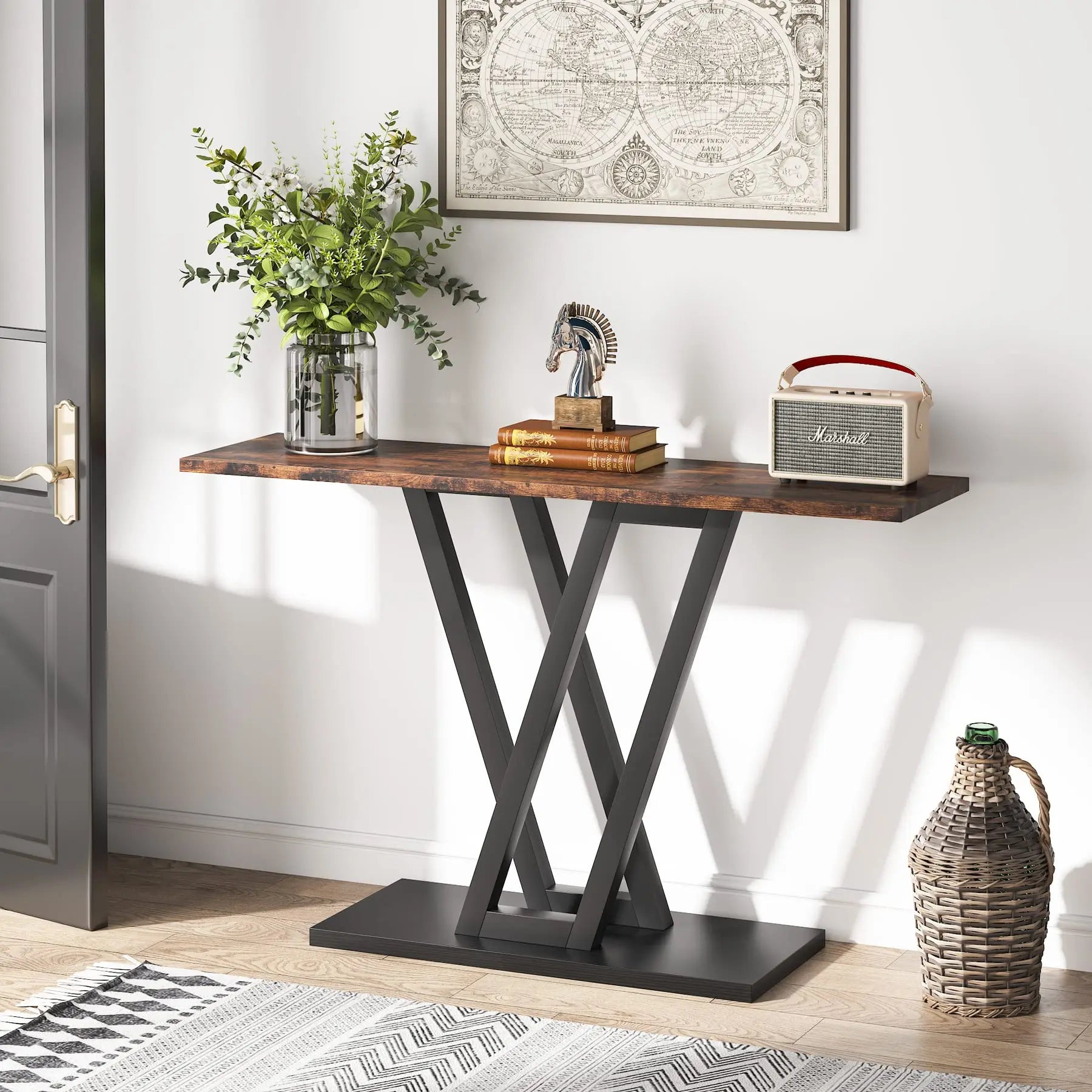 The Bradford Entryway Table by Morsale features a wooden top and sleek black crossed legs. It showcases a vase with a plant, a decorative sculpture, and a vintage radio beneath world map wall art. A wicker basket is placed on the floor nearby against the white wall, completing the living room setup.
