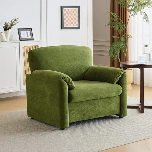 An Olive Green 38" Oversized Accent Chair by Morsale rests on a light gray rug in a cozy living area. Nearby, a small round wooden table holds a glass object. The backdrop features a white cabinet, framed art, and potted plants by a window with brown curtains.
