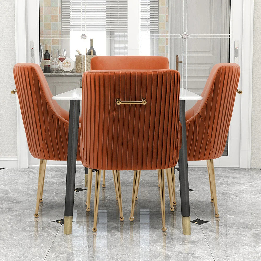 A dining room showcases a modern table with Morsale's Modern Glam Boucle Pleated Velvet Dining Chair Set of 4, featuring luxurious orange velvet upholstery and gold legs. Behind it sits a glass door with curtains, next to a small shelf displaying wine bottles and glasses on chic gray marble flooring.