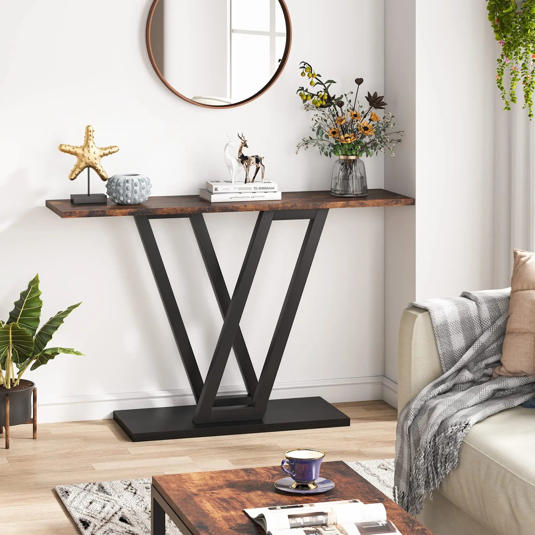 A modern living room showcases the stylish Morsale Bradford Entryway Table with black metal V-shaped legs, displaying a decorative starfish, books, deer figurine, and vase. A round mirror hangs above, accompanied by a coffee table, lush plant, comfy couch, and hanging greenery.