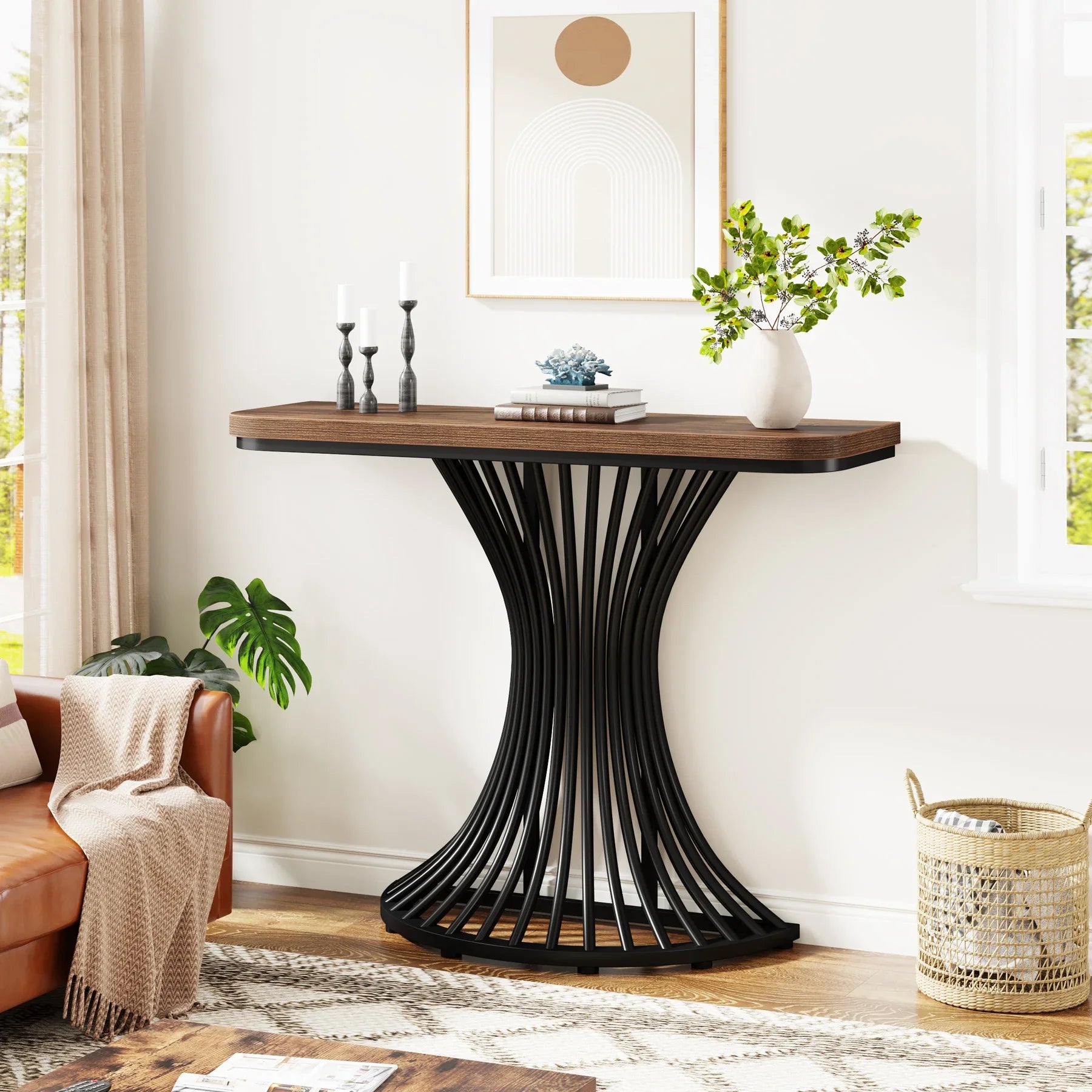A modern living room features the Hinton 39-Inch Entryway Table by Morsale. On it, chemical-free candles, books, and a plant add charm. Artwork enhances the setting while a floor potted plant sits beside a woven basket and a brown sofa with a black metal base providing stylish support.