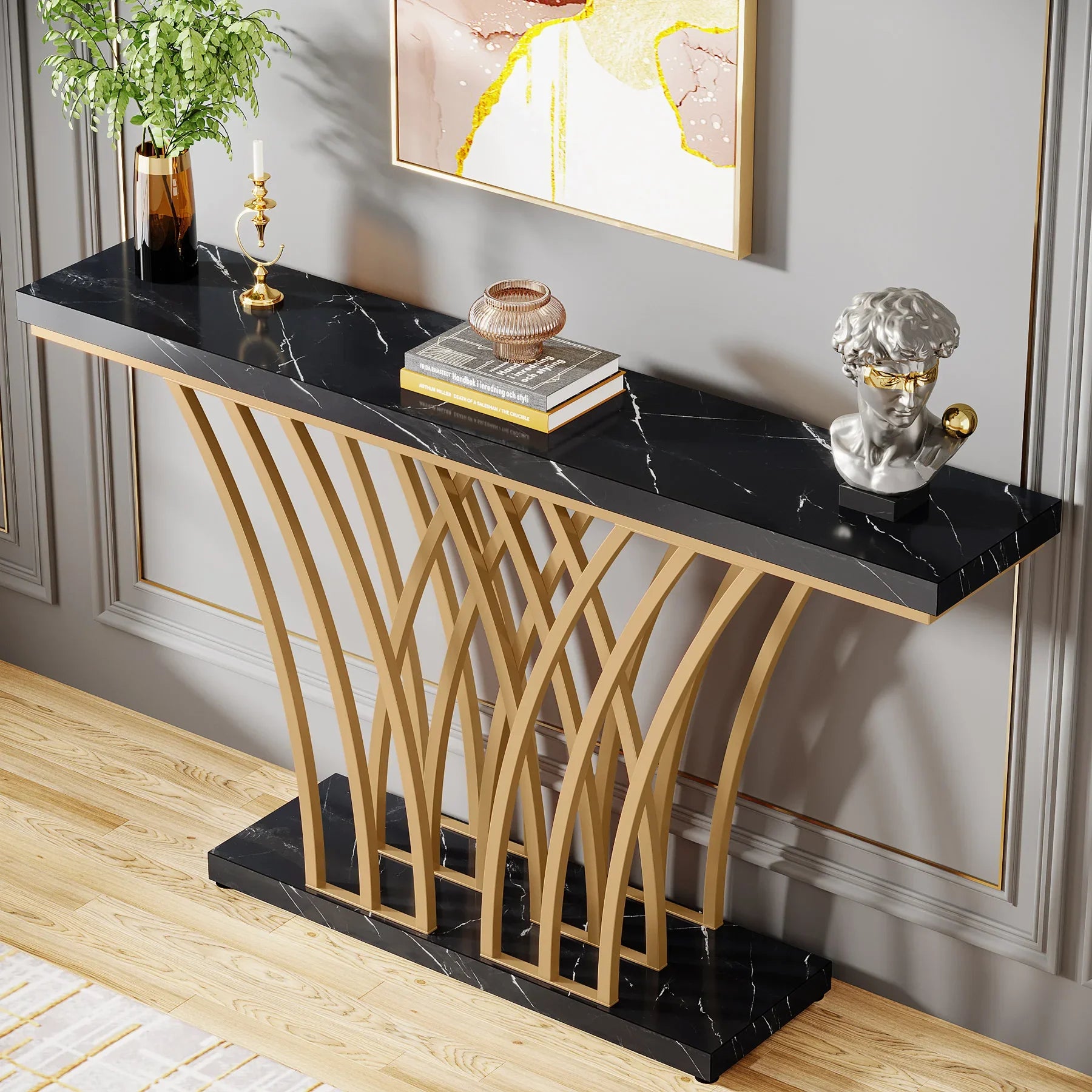 The Bassett Entryway Table by Morsale, with a black faux marble finish and geometric gold metal base, stands against a gray wall holding books and decor. Above it, an abstract framed artwork complements the light wooden floor beneath.