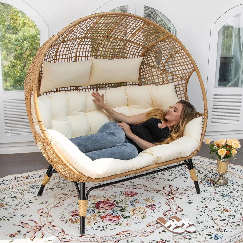 A woman unwinds on the Morsale Winsford Patio Lounge Chair, laptop in hand, surrounded by lush greenery. A patterned rug and flowers adorn the foreground. This modern, cushioned outdoor furniture provides a sunny and serene ambiance ideal for leisurely afternoons.