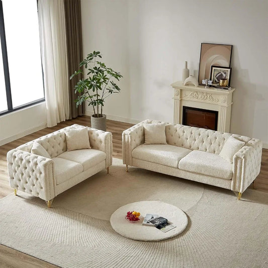 The living room showcases a Morsale 2-Piece Loveseat Sofa Set in cream chenille upholstery on a light rug, accompanied by a round coffee table with books and flowers. A potted plant sits by a large window, while framed art and candles grace the small fireplace, enhancing modern elegance.