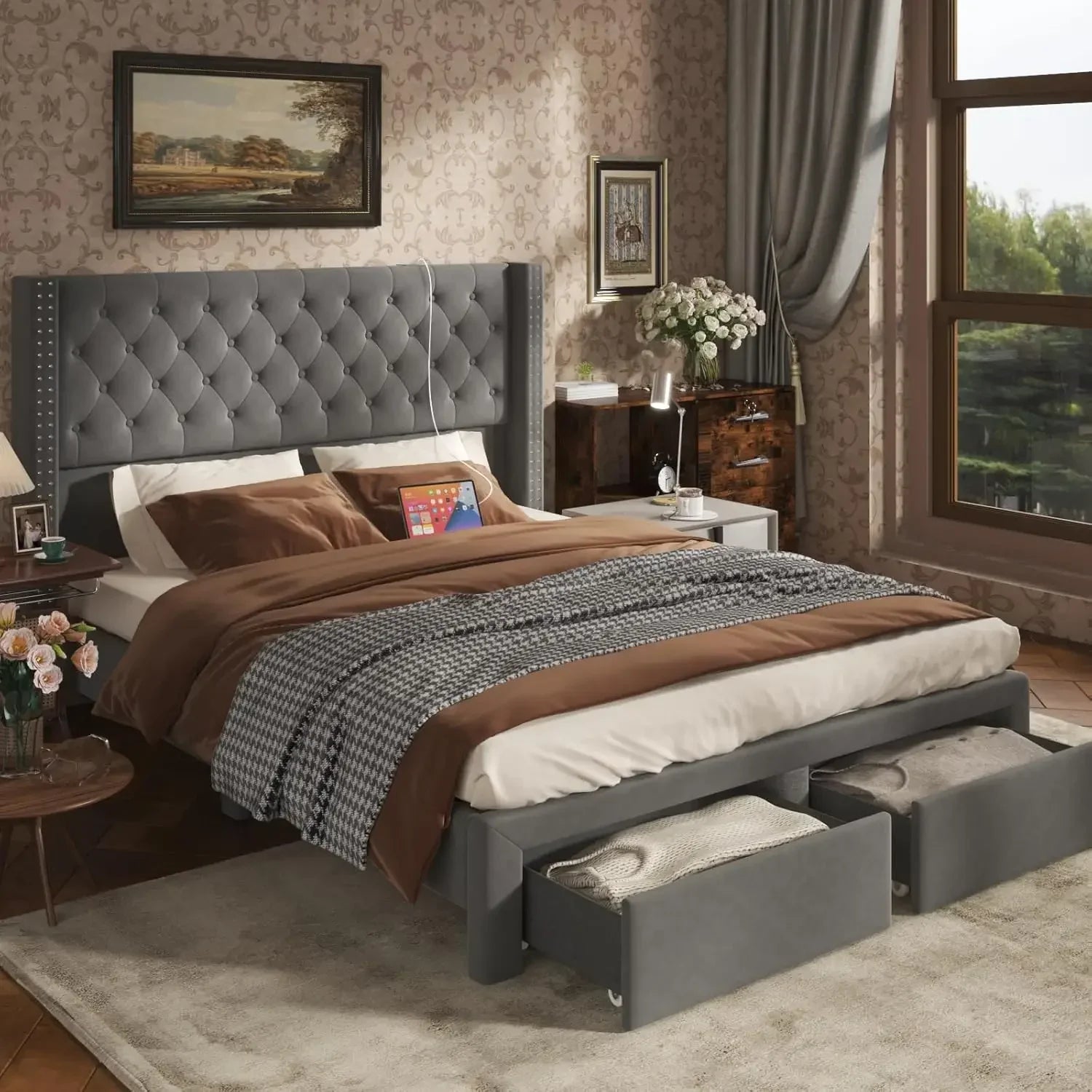 A bedroom showcases the Morsale Gio Velvet Queen Bed with Storage, velvet-upholstered padded headboard, open base drawers, patterned bedding, side tables with charging ports, lamps, flowers, wall artwork, a large window with curtains, and a patterned rug.