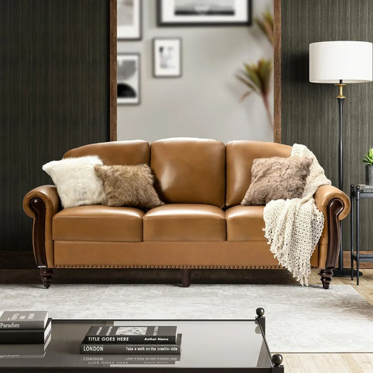 The Modi Genuine Leather Sofa 3-Seater Couch by Morsale, in brown with elegant nailhead trim, is in a chic living room. It features three cushions—white, brown, and gray—and a cream throw blanket on one armrest. A floor lamp and framed pictures enhance the dark wall.