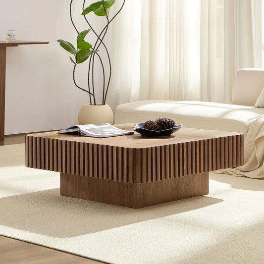 A stylish living room displays the Morsale Belford Walnut Coffee Table, featuring vertical grooves, with an open book and decorative item on top. A white sofa and potted plant sit in the modern setting as natural light filters through sheer curtains.