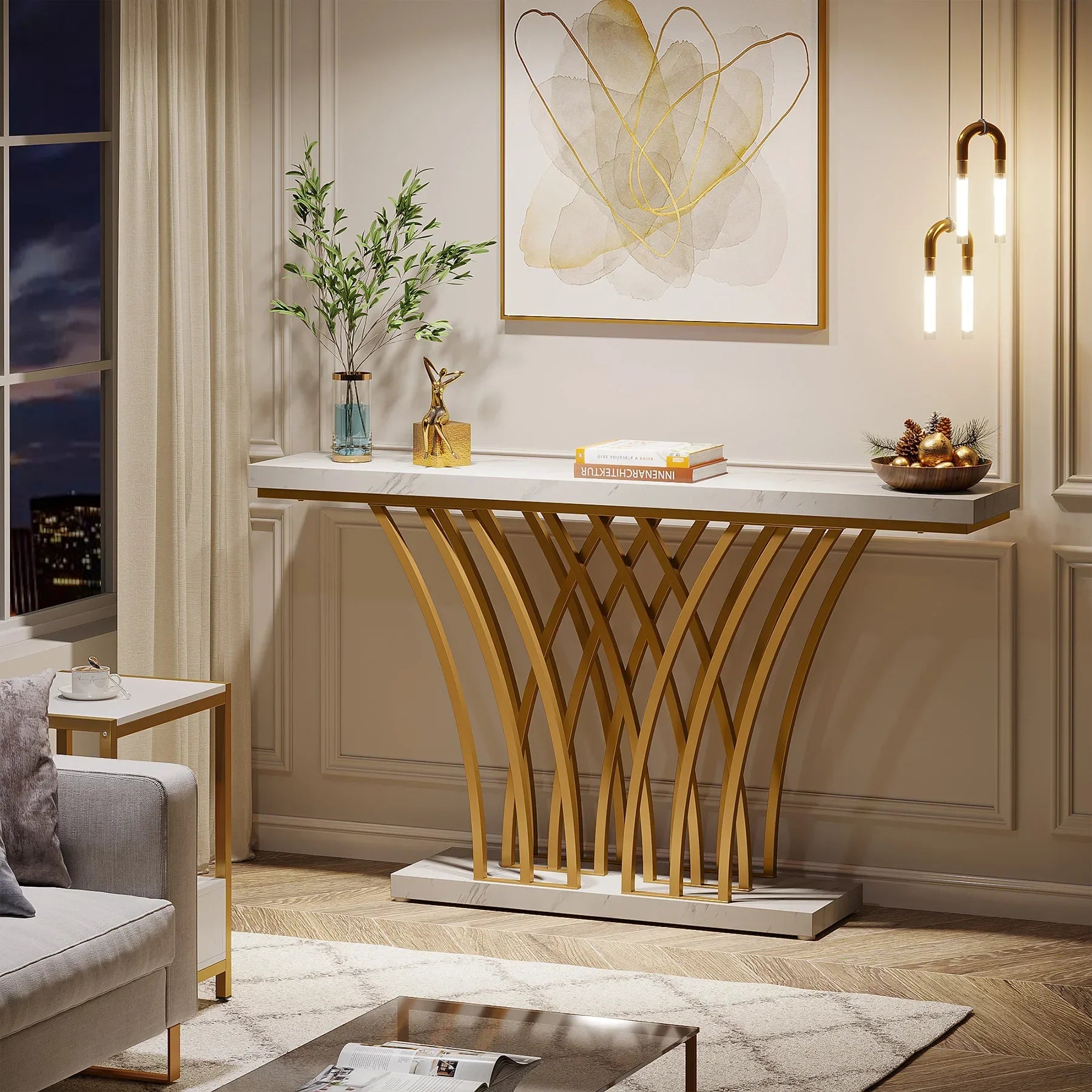 A luxurious living room corner features the Morsale Bassett Entryway Table, boasting a modern design with a wavy gold base and marble top. Contemporary art, hanging lights, a plant, and books complement the decor, while a cozy sofa and night view complete this exquisite setting.