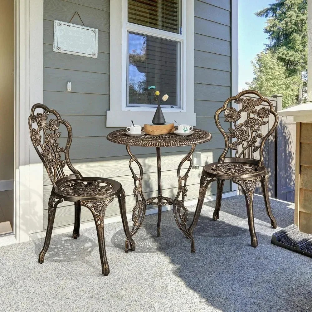 A small outdoor patio showcases the Morsale Davids 3-Piece Vintage Outdoor Set—an ornate metal table effortlessly assembled with two matching chairs. The table accommodates a teapot, two teacups, and a book. In the surroundings are a part of a house wall and glimpses of trees.