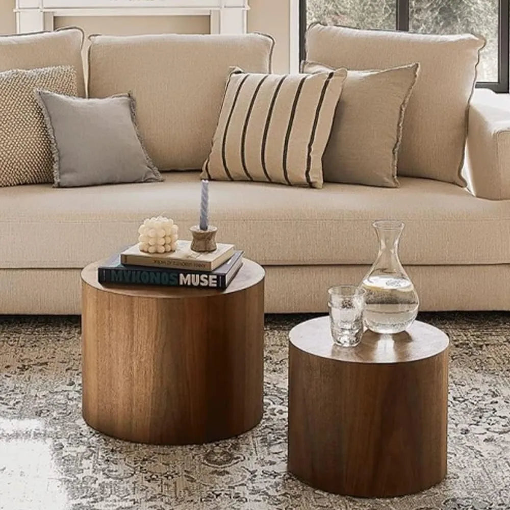 A cozy living room features a beige couch with pillows, and in front, the Morsale Living Room Wood Coffee Tables (Set of 2) hold a book, candle, and glass carafe. A textured rug lies underneath as light filters through the window, highlighting the elegant setup.