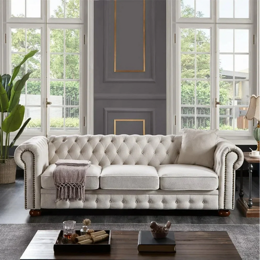 Luxurious living room showcasing a modern beige 3-Seat Chesterfield Sofa by Morsale, accessorized with cushions and a throw. Positioned against grand windows overlooking greenery, the space features elegant wall paneling and a dark wood coffee table with decor, adding warmth and sophistication.