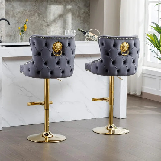 The Morsale Pull Ring Velvet Barstools Set of 2, with tufted gray upholstery and golden lion head accents, sit at a white marble counter. Featuring chrome frames and adjustable height, the stools enhance the wooden floor, large windows, and potted plant. A bottle and glasses await on the counter.