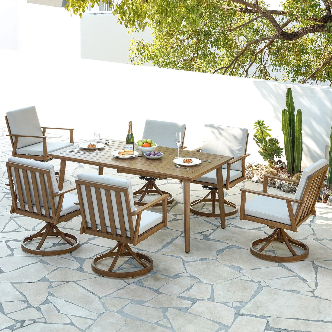 A modern patio scene showcases the Morsale Shefford 7-Piece Patio Dining Set with a wooden table and six cushioned chairs. The table is adorned with plates, a champagne bottle, glasses, and a fruit bowl. A large tree and cactus are in the background on the stone patio.