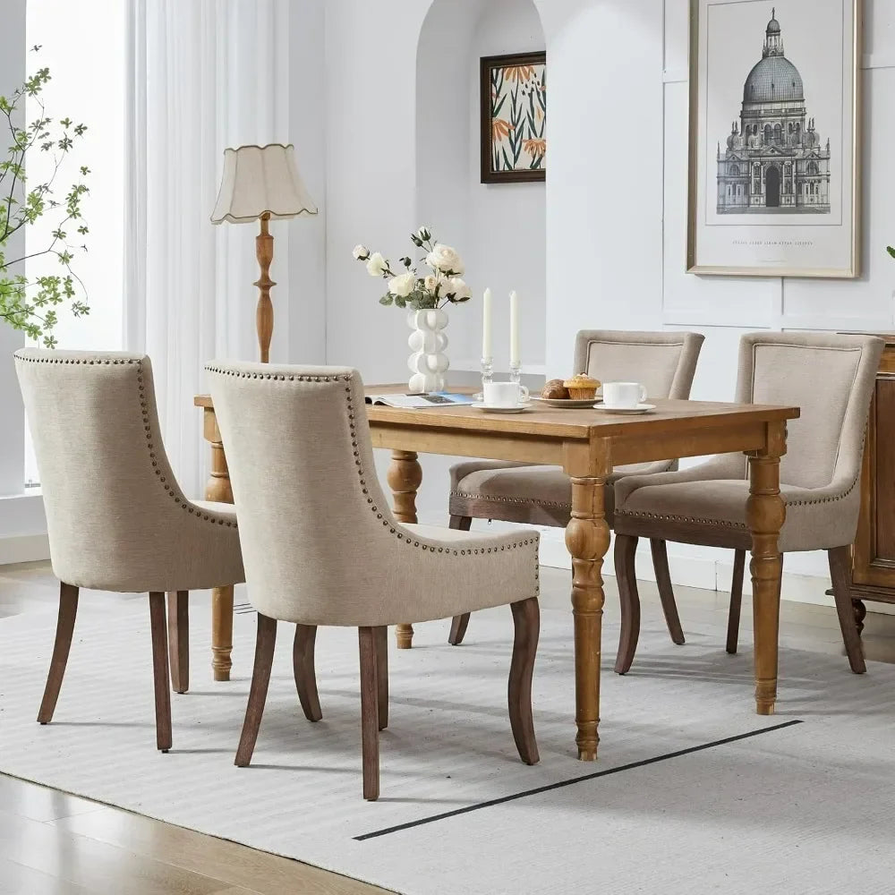A modern dining room showcases a sleek wooden table with six Verona dining chairs by Morsale in beige upholstery. The table features candles, flowers, and a book, complemented by a lamp and framed art. Light filters through sheer curtains, highlighting the lush greenery outside.