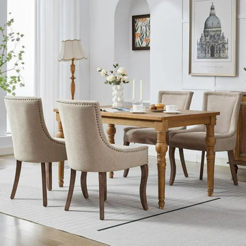 A modern dining room showcases a sleek wooden table with six Verona dining chairs by Morsale in beige upholstery. The table features candles, flowers, and a book, complemented by a lamp and framed art. Light filters through sheer curtains, highlighting the lush greenery outside.
