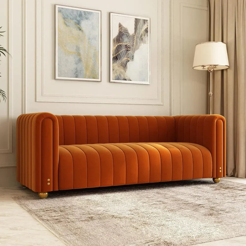 A modern living room features Morsale's 80" Velvet Living Room Sofa in orange with vertical tufting and round gold legs. Two abstract paintings hang above, while a beige textured rug covers the floor, accompanied by a tall floor lamp to the right.
