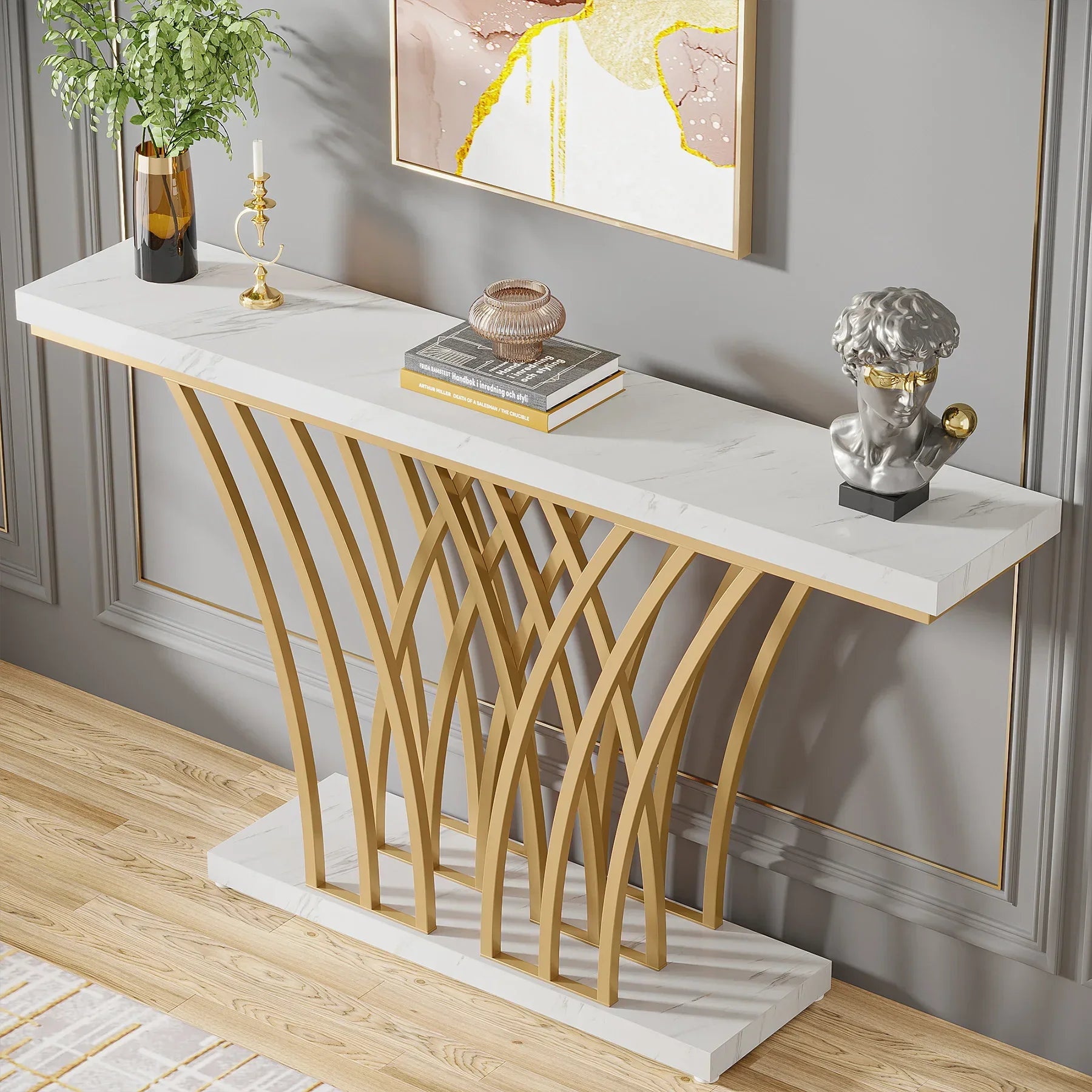 The Bassett Entryway Table by Morsale features a white marble top and gold geometric legs. A stack of books, bust sculpture, decorative jar, candleholder, and leafy plant adorn it. A contemporary art piece graces the wall behind while the light wood floor enhances this elegant furniture piece.