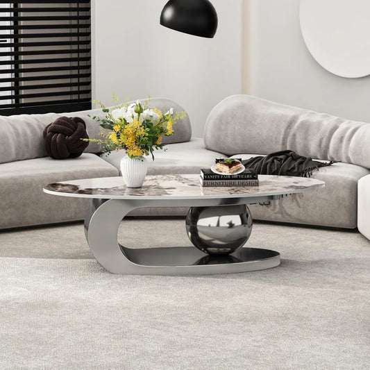 A modern living room showcases a light gray curved sectional sofa, reflecting Morsale's style commitment. An oval coffee table with a ceramic top and steel base sits on a gray rug, adorned with chemical-free yellow flowers. On the table rests "Vanity Theaters.