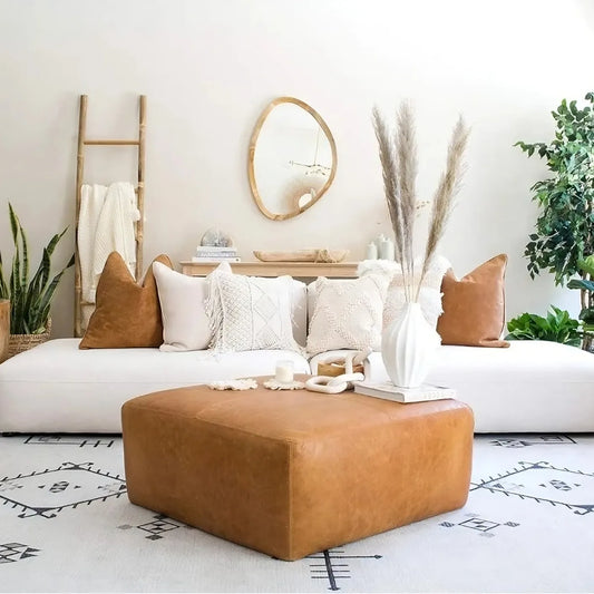 A cozy living room showcases a white US Origin sofa with beige and tan cushions, a Morsale Leather Ottoman in Cognac Tan on a patterned rug, and wall decor featuring a round mirror and ladder with throw blanket. Potted plants and chemical-free pampas grass in a vase complete the scene.